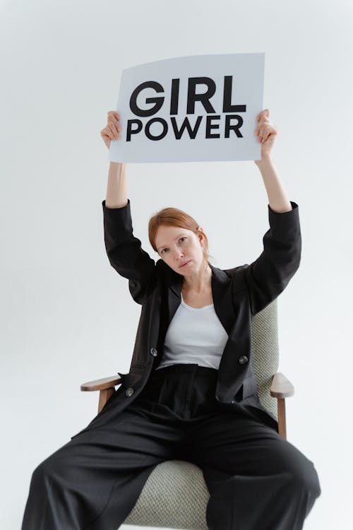 A Woman in Black Blazer and Pants Sitting on the Chair while Holding a Placard