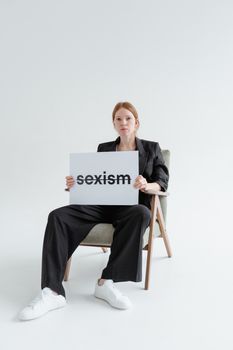 Woman in black corporate attire holds sign discussing sexism, sitting on chair with white background.
