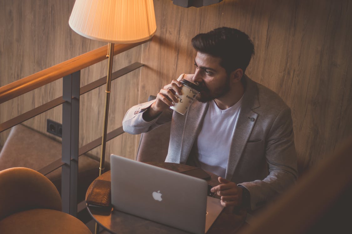 A Man in Gray Suit Drinking Coffee while Sitting Near His Laptop on the Table