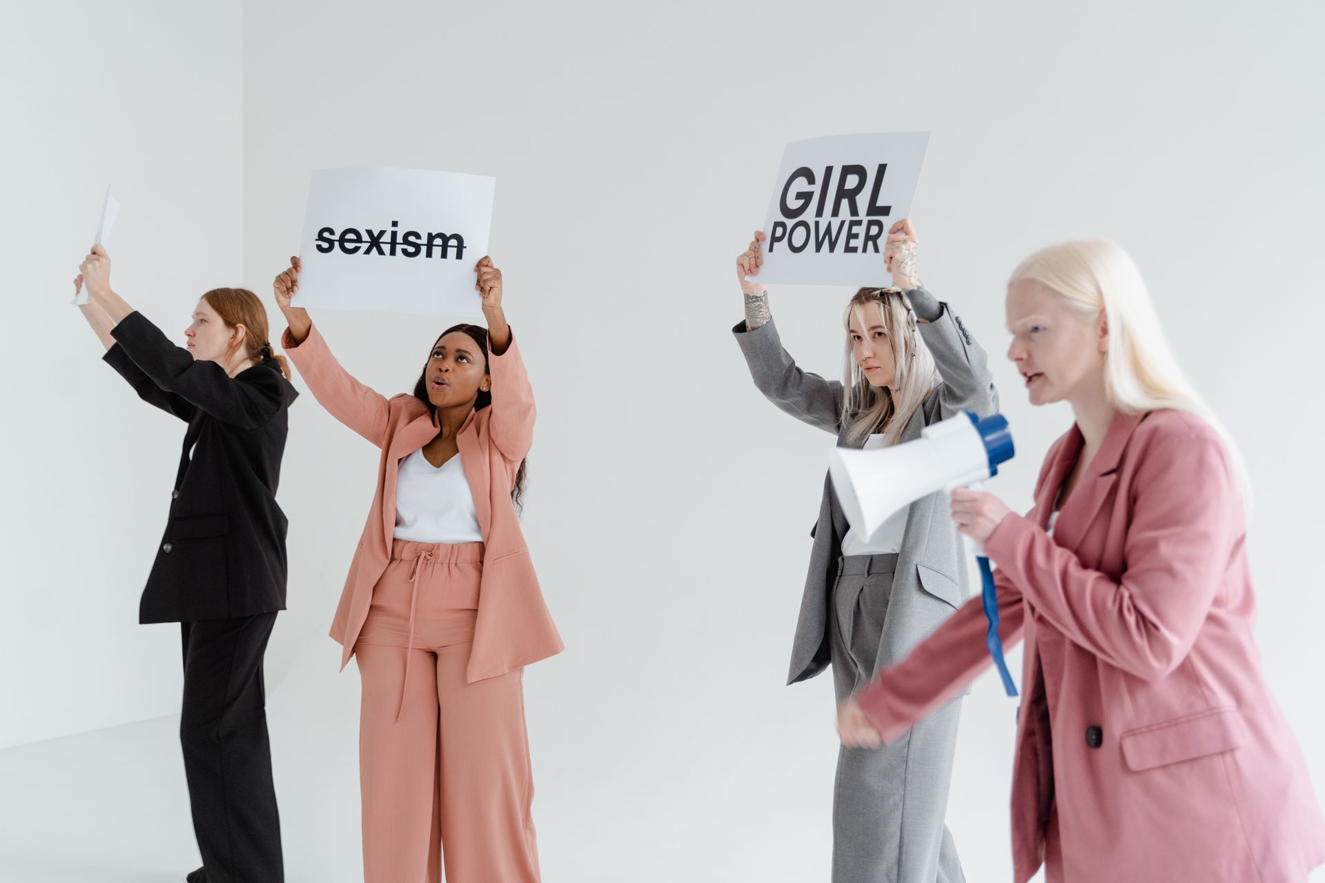 Four women in business attire protest with placards against sexism on a white background.