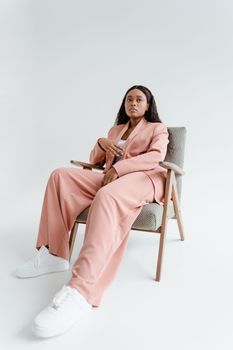 Black woman in a pink suit sitting on a chair, embracing fashion in a studio setting with a white background.