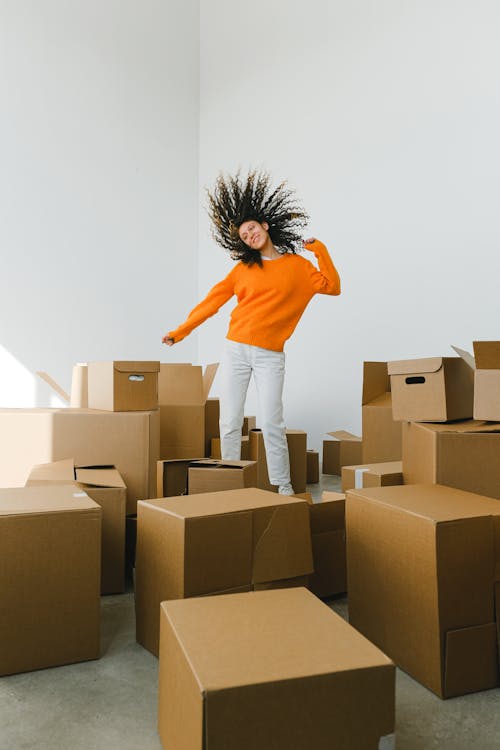 Joyful female with moving hair dancing among piles of carton containers after renting house on light background