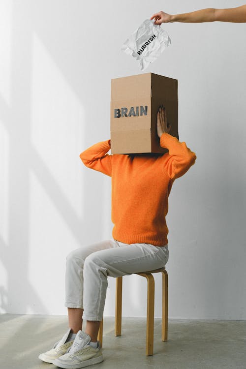 Free Crop anonymous person putting creased paper with Rubbish inscription in container with Brain title on head of female on chair Stock Photo