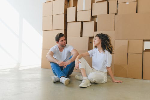 Woman talking to boyfriend with crossed legs sitting on floor against piles of carton containers after moving into house