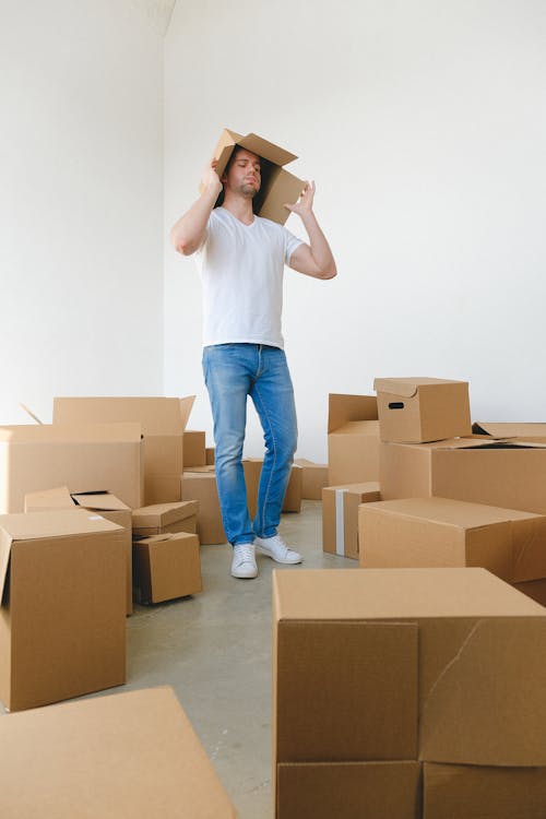 Male with cardboard box on head between rows of belongings in new house on light background