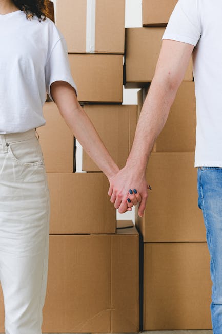 Crop unrecognizable couple in casual clothes holding hands while standing near pile of carton boxes after moving into new apartment