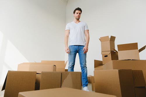 Low angle of serious young guy with dark hair in casual outfit standing near pile of cardboard boxes in light room and looking away while preparing for relocation
