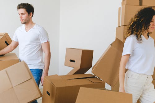 Serious young man and woman in white t shirts carrying carton boxes in light room while moving into new apartment together