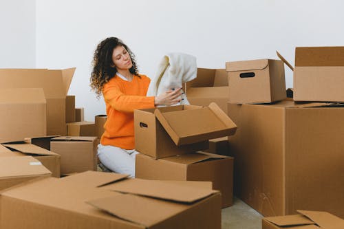 Calm young relocating woman with curly dark hair in stylish sweater packing belongings into pile of cardboard boxes sitting on floor before moving into new apartment