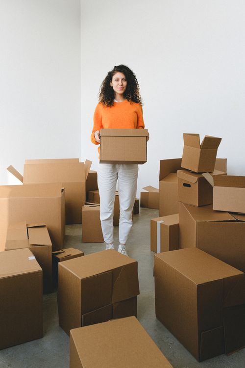 Free Full body of young female with dark curly hair carrying cardboard boxes and looking at camera while relocating to new apartment Stock Photo