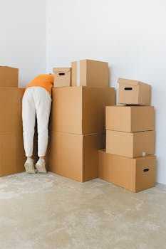 Back view of unrecognizable female in casual clothes arranging various cardboard boxes near white wall after moving into new empty apartment in daylight