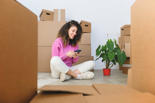 Delighted young female homeowner sitting near pile of boxes and browsing smartphone