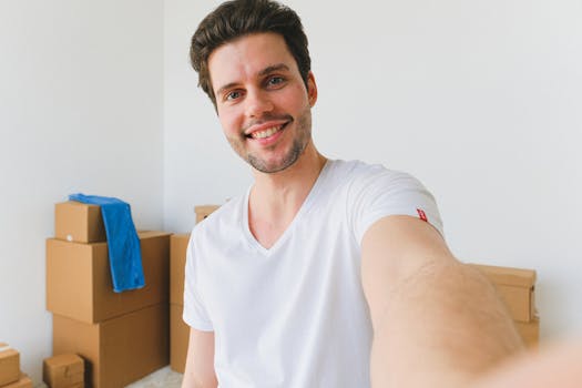 Delighted young relocating man with dark hair in white t shirt smiling and looking at camera while taking selfie against pile of cardboard boxes in new purchased apartment