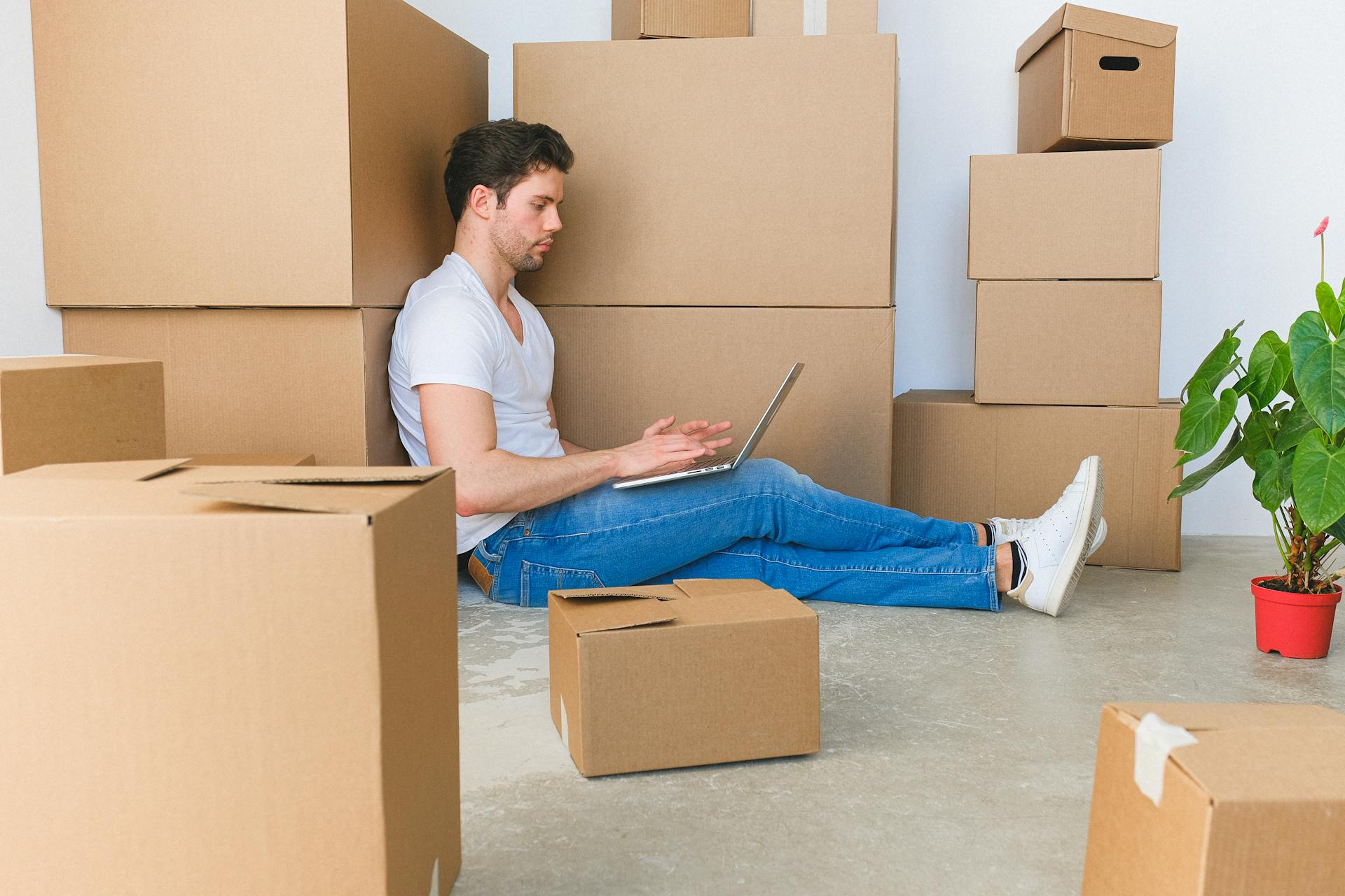 A man seated on the floor using a laptop, surrounded by cardboard boxes in a moving scenario.