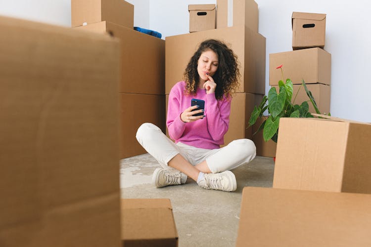 Excited Young Woman Messaging On Smartphone Sitting On Floor Near Boxes After Relocation