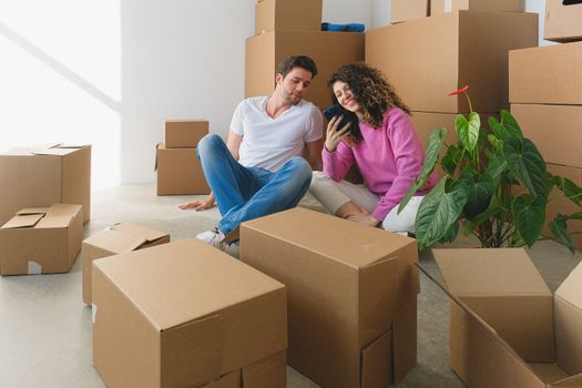 Young couple sitting among cardboard boxes, looking at smartphone, symbolizing moving or relocation.
