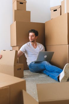 Young focused male freelancer checking items in carton box while sitting on floor and working online on laptop before moving into new flat