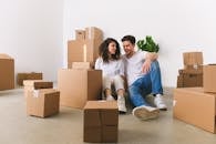 Full body of cheerful couple talking while resting on floor near belongings with carton boxes after moving into new apartment