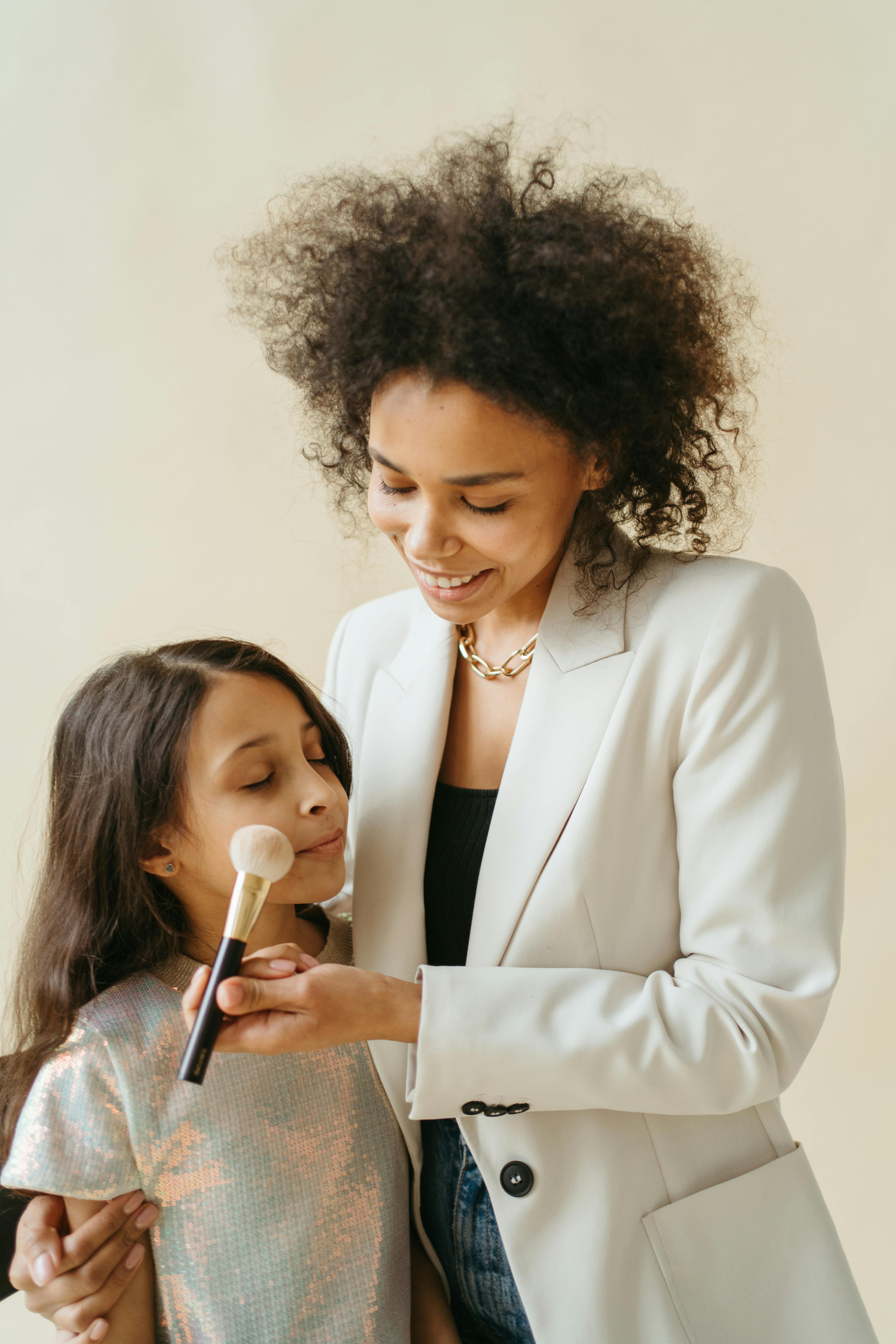 a mother putting a makeup on her daughter s face while holding a brush