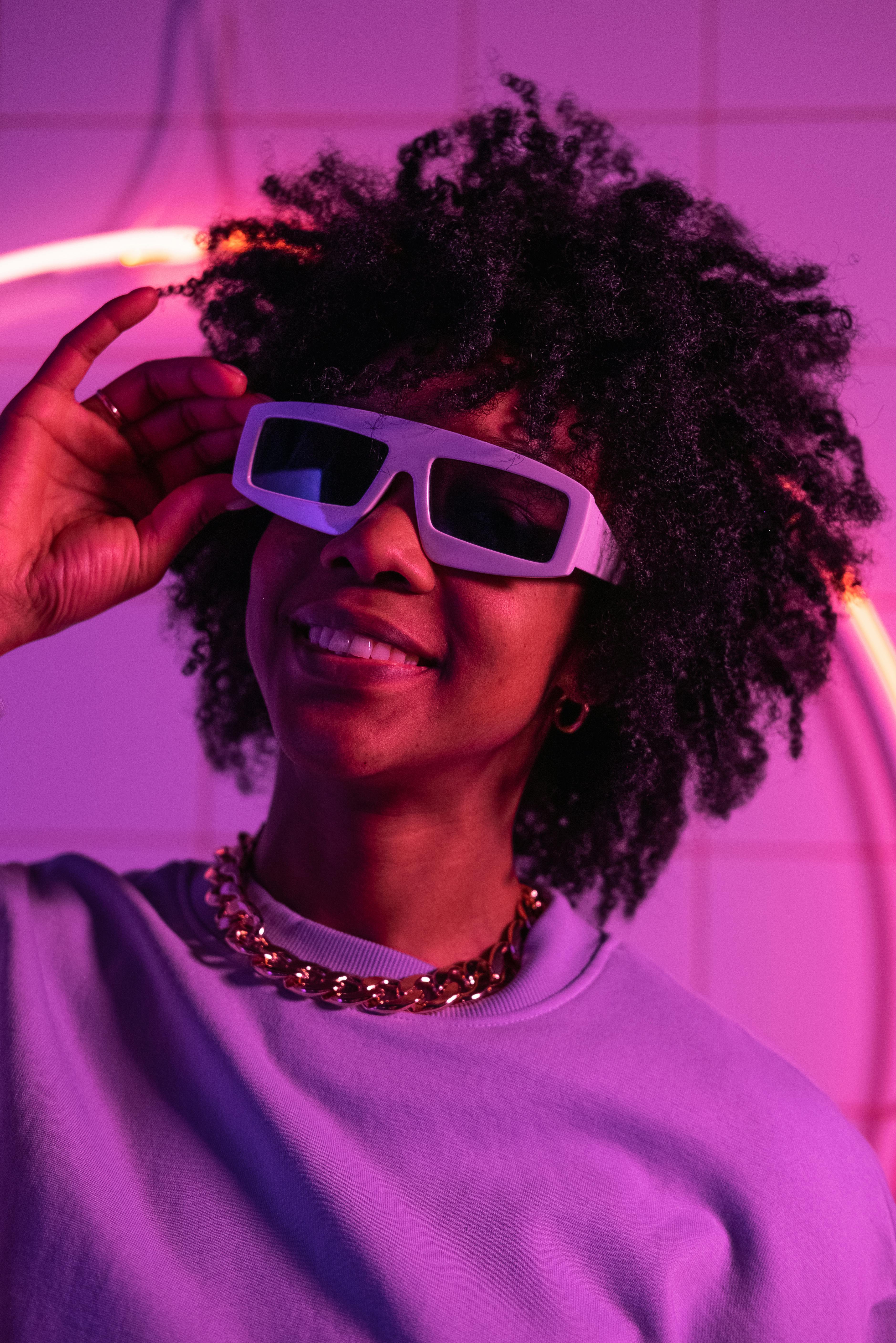 Stylish portrait of a smiling Black woman with afro hair and sunglasses under neon lights.