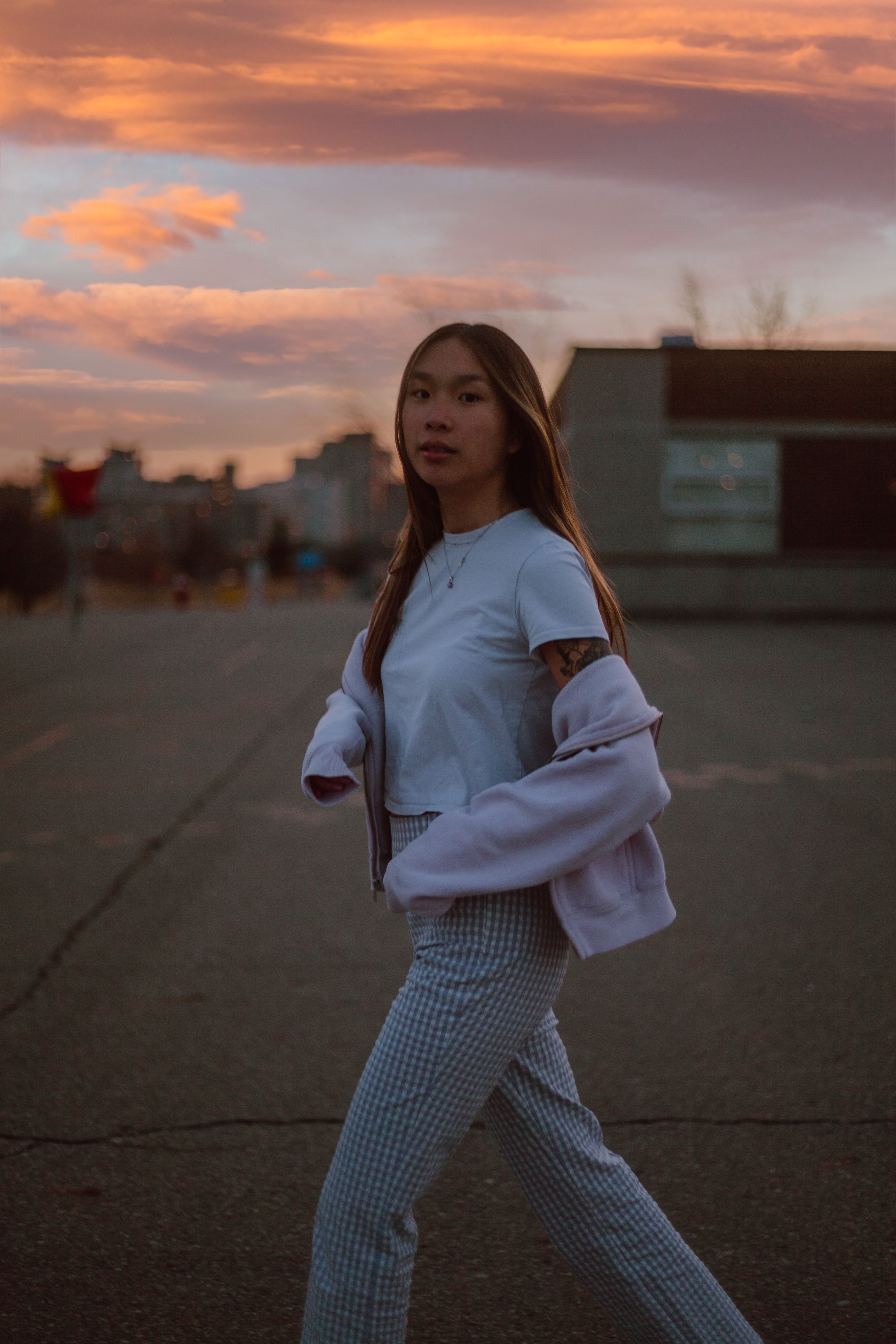 Asian woman walks confidently outdoors against a vibrant sunset backdrop in the city.
