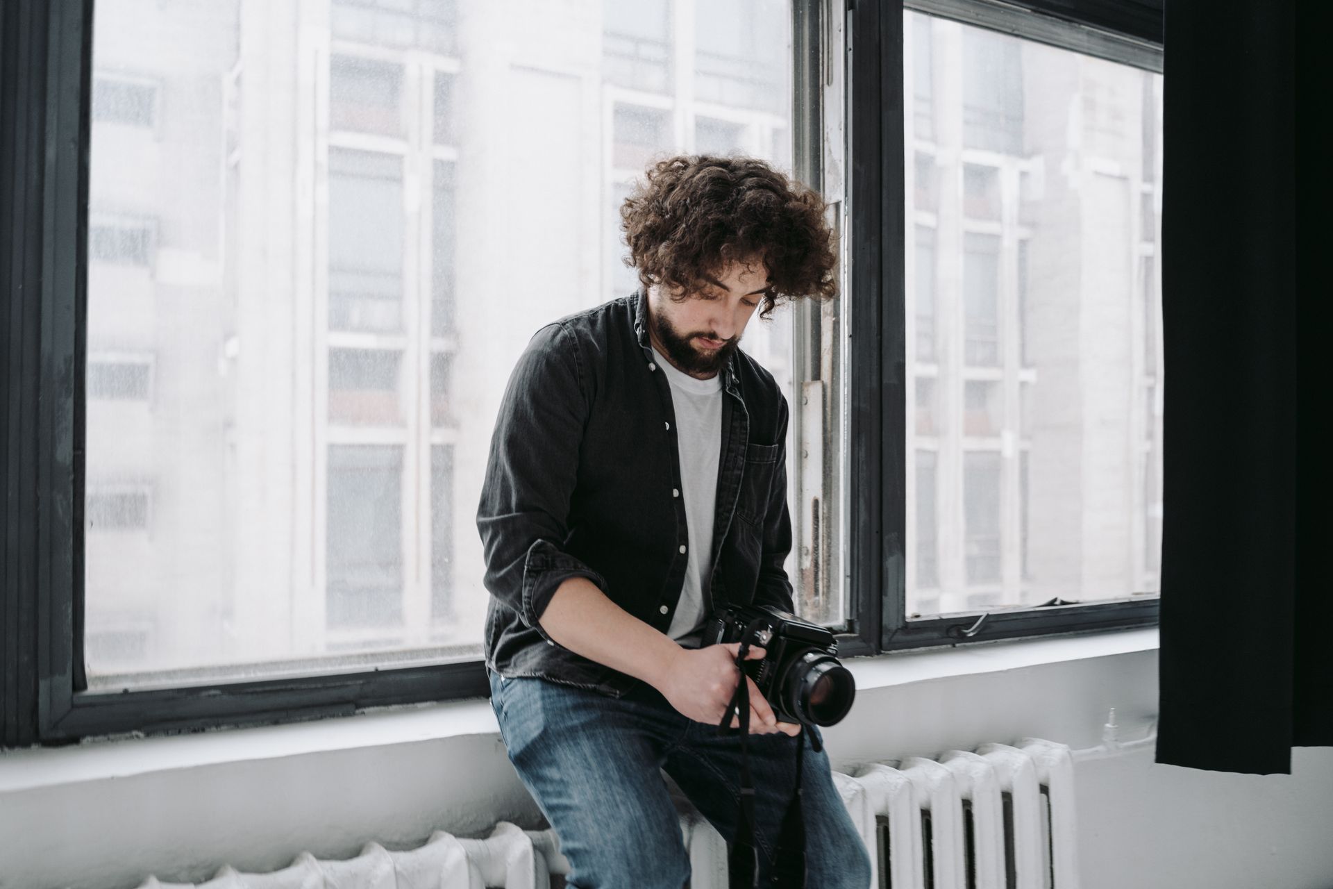 A contemplative male photographer sitting indoors by a large window holding a camera.