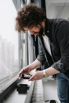 A man carefully loads film into a camera next to a large window with natural light.