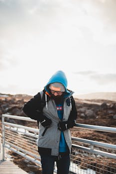 Young male in warm clothes standing on viewpoint near white fence on hill under cloudy sky in winter