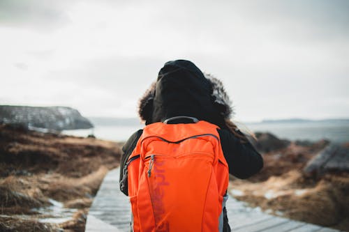 Back view of unrecognizable tourist in outerwear with bright orange backpack standing on wooden walkway on sea shore in winter