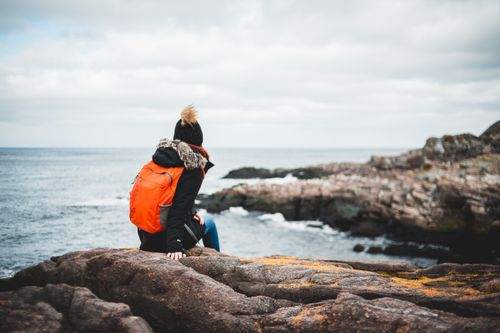 Free Back view of anonymous female hiker in warm jacket and hat with orange backpack sitting on rocky cliff and admiring wavy ocean against cloudy sky Stock Photo