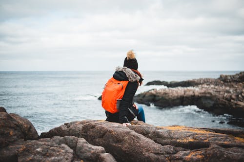 Back view of unrecognizable female traveler in warm clothes and backpack resting on rough rocky cliff and admiring stormy sea under cloudy sky