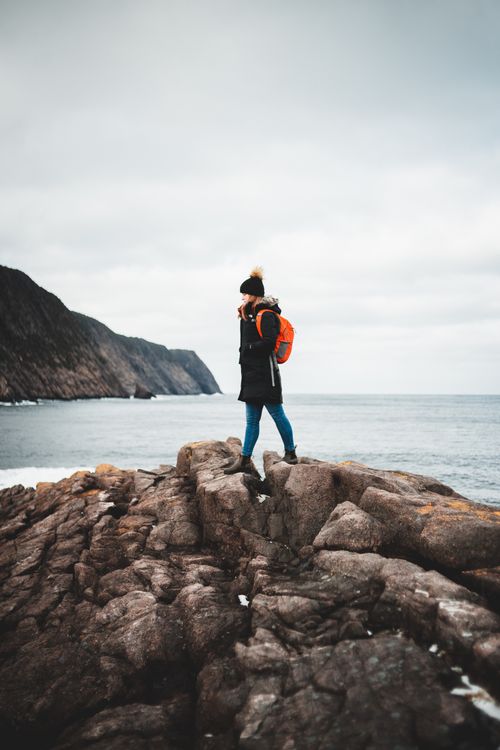 Free Side view of young female hiker in warm jacket and hat walking on massive rocky cliff and observing seashore under overcast sky Stock Photo