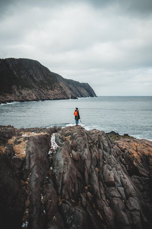 Back view of distant unrecognizable female hiker in warm clothes with bright orange backpack recreating on rocky cliff and admiring picturesque scenery of ocean under gloomy sky