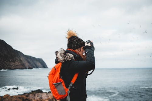 Side view of young female photographer in warm jacket and hat with bright orange backpack taking pictures of powerful stormy sea while standing on rocky shore during hiking trip