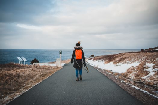 Back view of unrecognizable female hiker in puffer jacket and hat with backpack and photo camera in hand walking along asphalt road toward wavy sea against cloudy sky