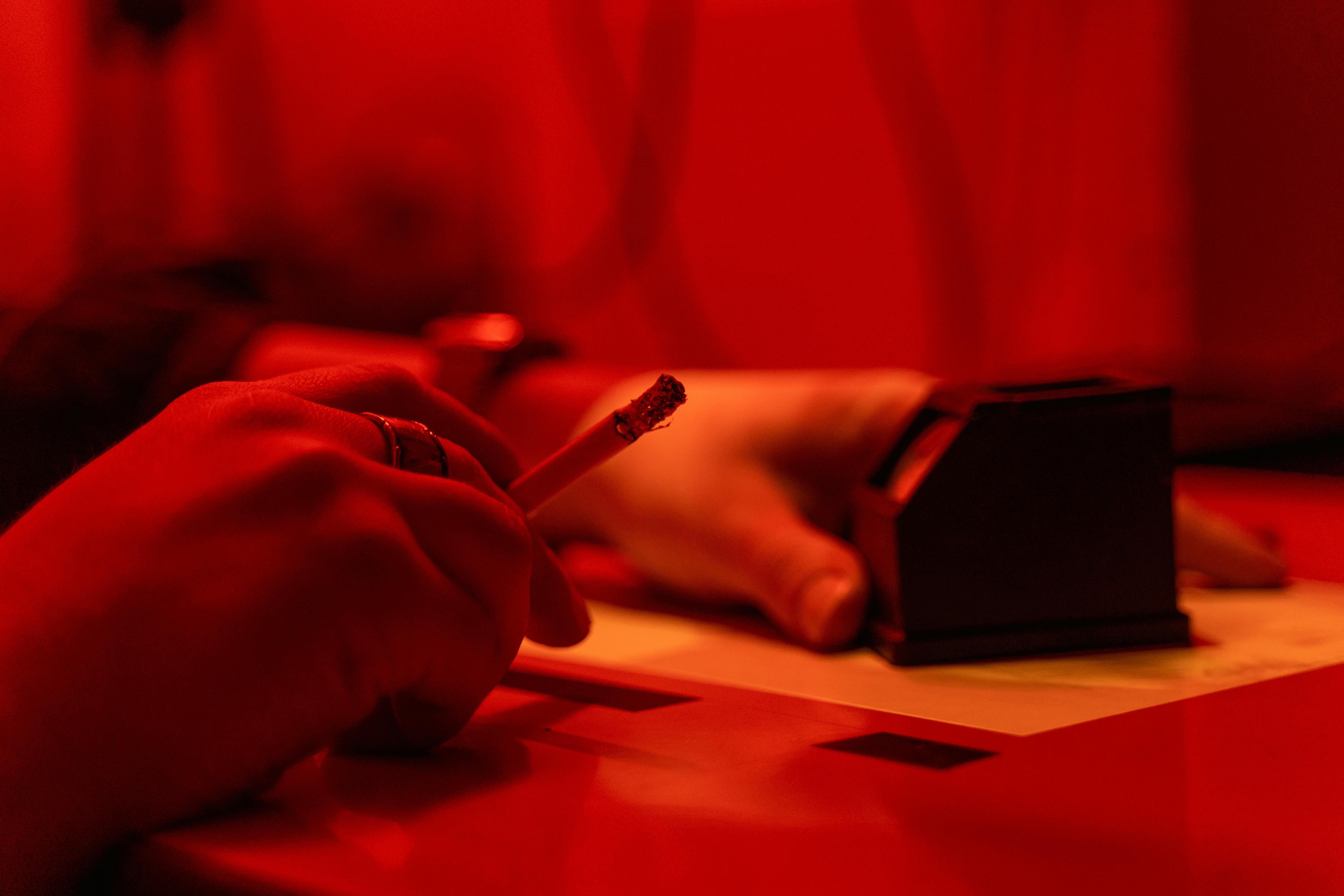 A hand holding a cigarette under ambient red lighting, creating a moody atmosphere.