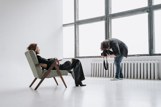 A photographer capturing a woman in a stylish chair during a minimalist indoor photoshoot.