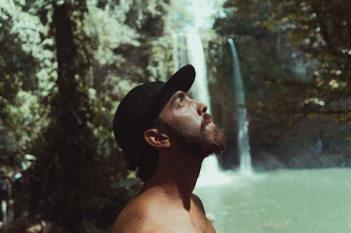 Close-Up Shot of a Bearded Man in Black Cap near a Waterfalls