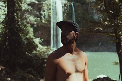 Close-Up Shot of a Shirtless Man in Black Cap Near a Waterfalls