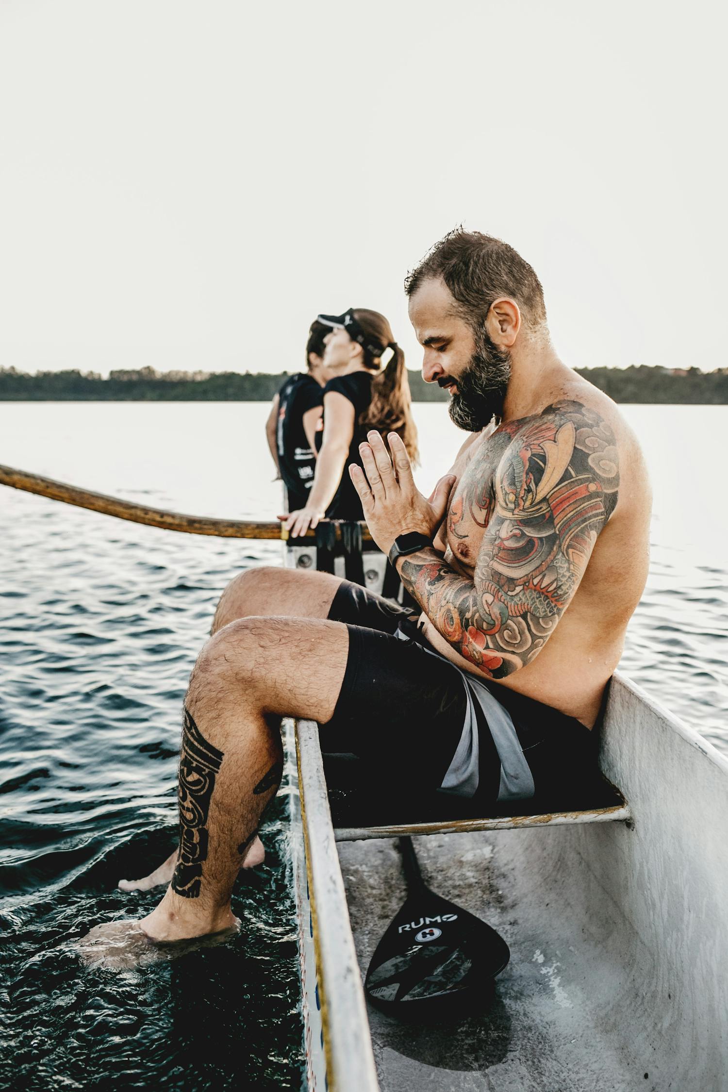 Tattooed sportsman praying in boat against unrecognizable friends on river