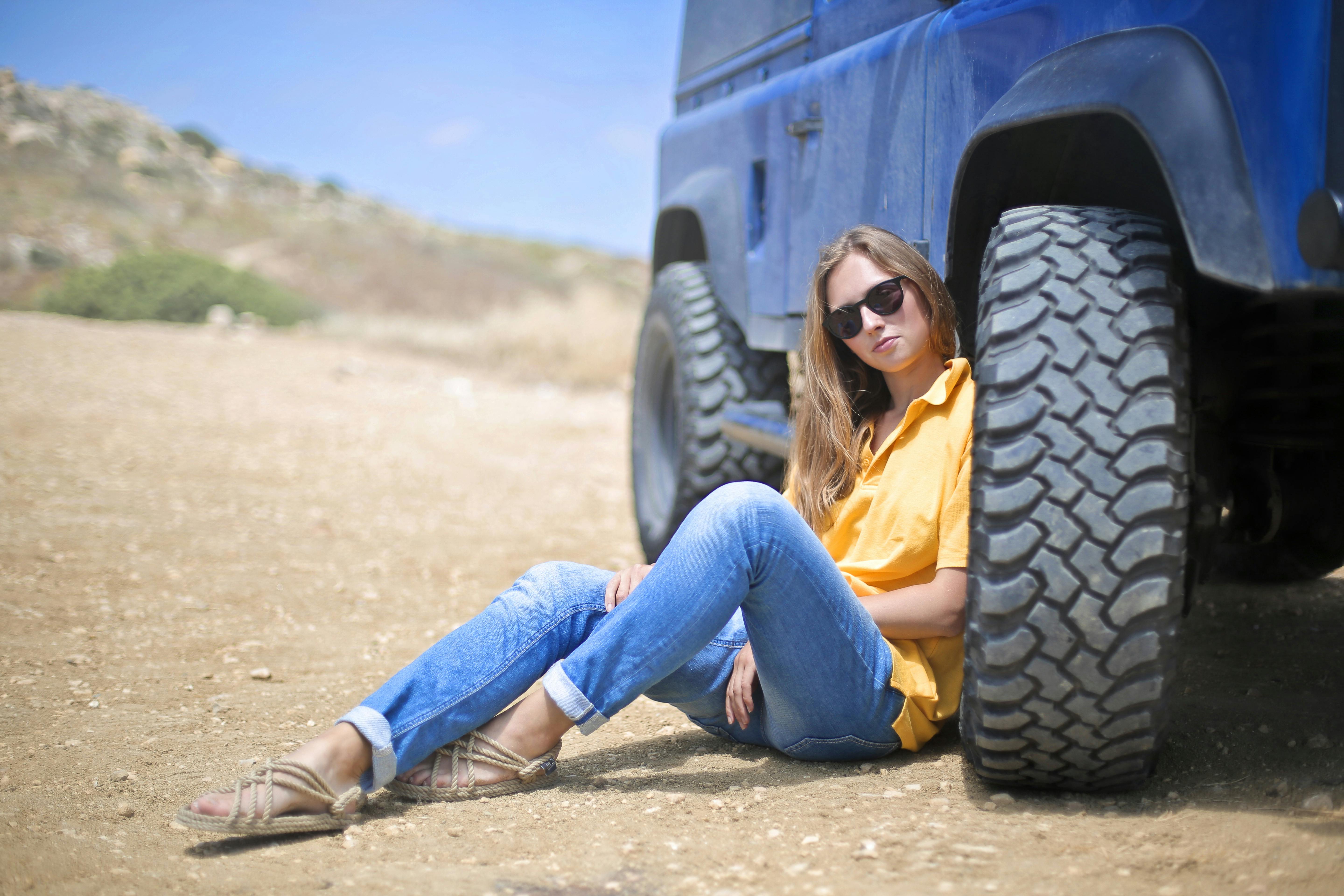 woman in yellow polo shirt sitting on ground leaning on blue vehicle at daytime