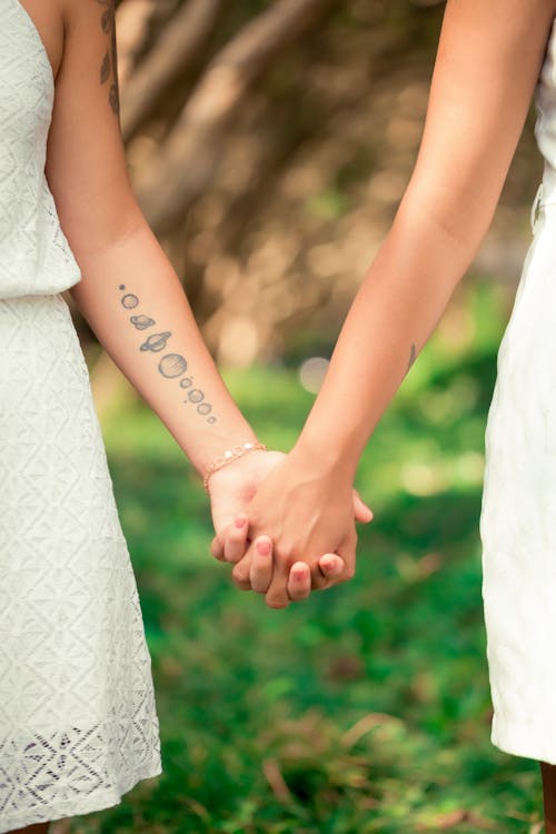 Crop anonymous young tattooed women holding hands gently on blurred background of fresh verdant grass in park