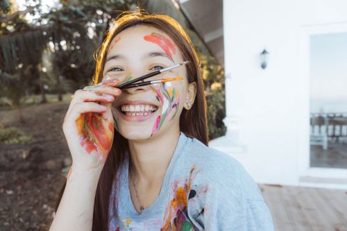 Free Smiling girl with paint all over her face and brushes held near her nose, enjoying outdoor creativity. Stock Photo