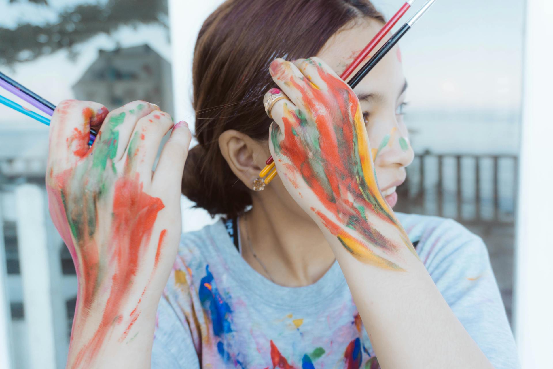 A young woman covered in colorful paint holds brushes against a blurred background.