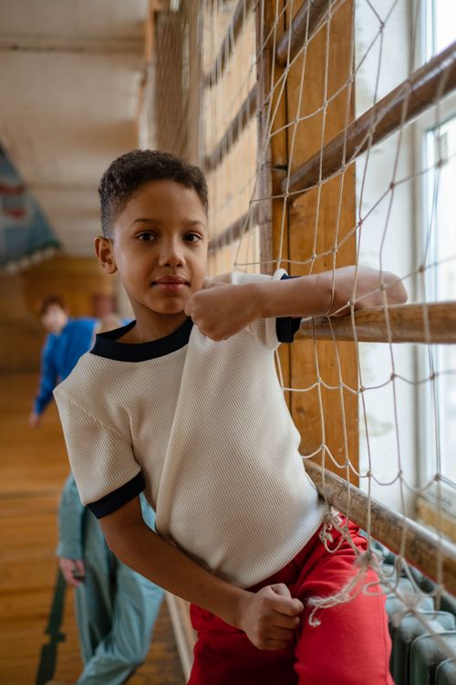 Free A young boy leans on a net in a gymnasium, wearing a white shirt and red pants. Stock Photo