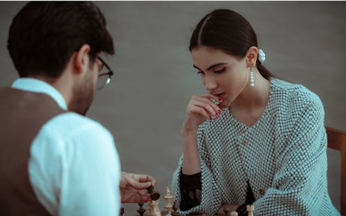 Focused ethnic female touching chin while thinking on chess tactic during play with male opponent at gray background