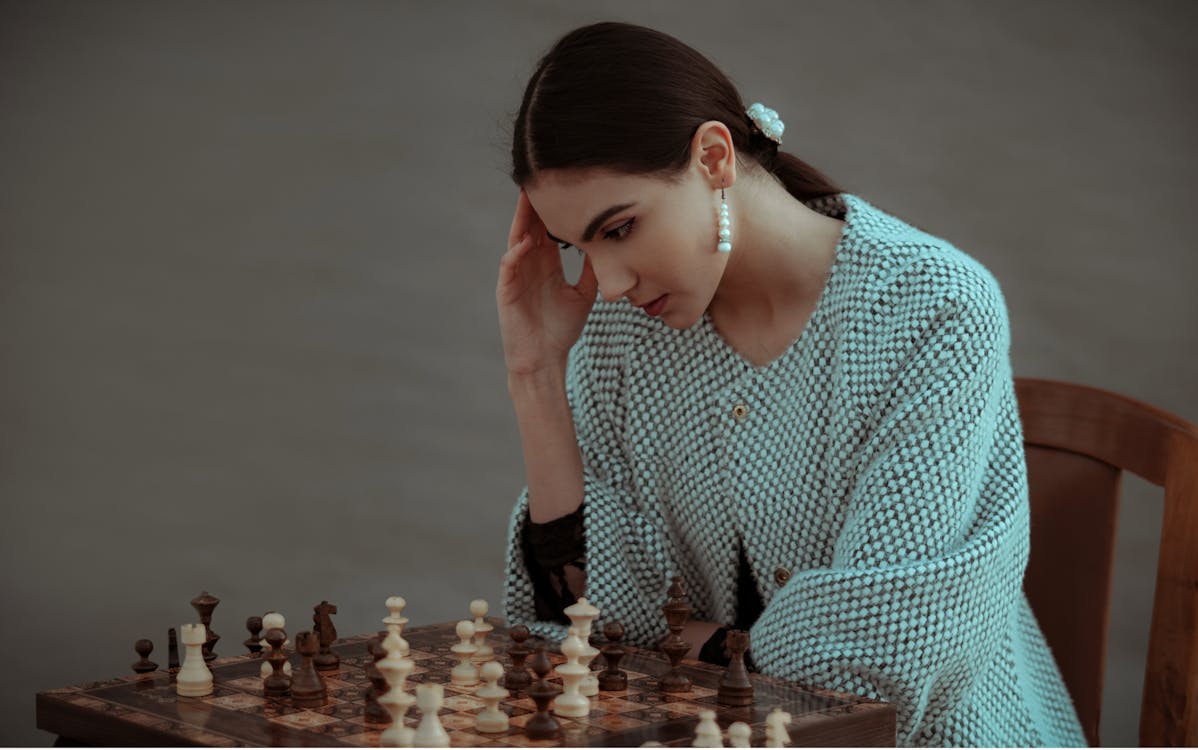 Concentrated ethnic female sitting on chair and touching head while playing chess against blurred gray background