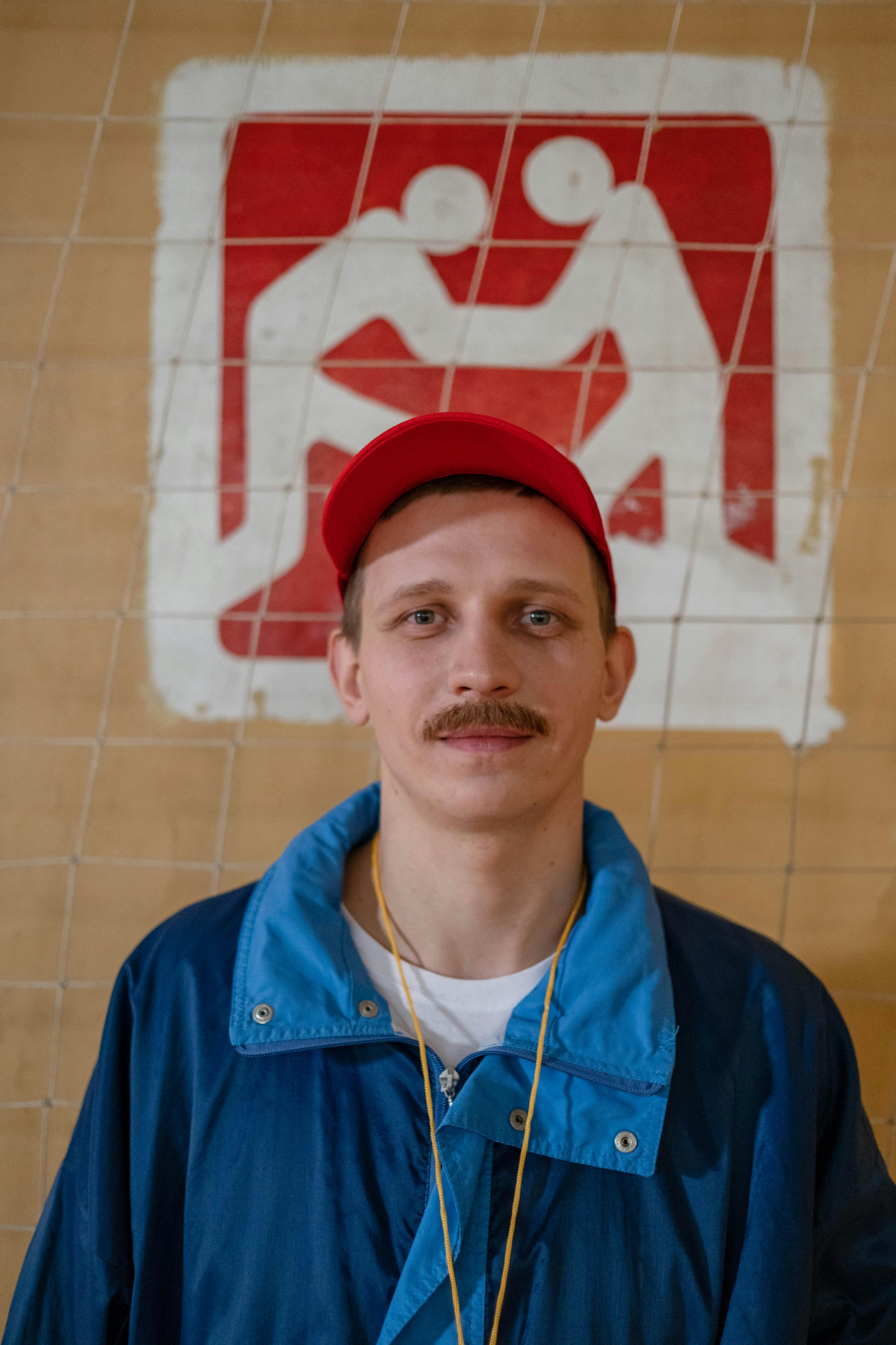 Image of a male sports coach with a moustache, wearing a blue jacket and red cap, standing indoors.