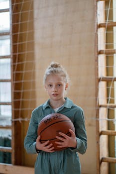 Caucasian girl holding basketball in indoor gym with netting in background.
