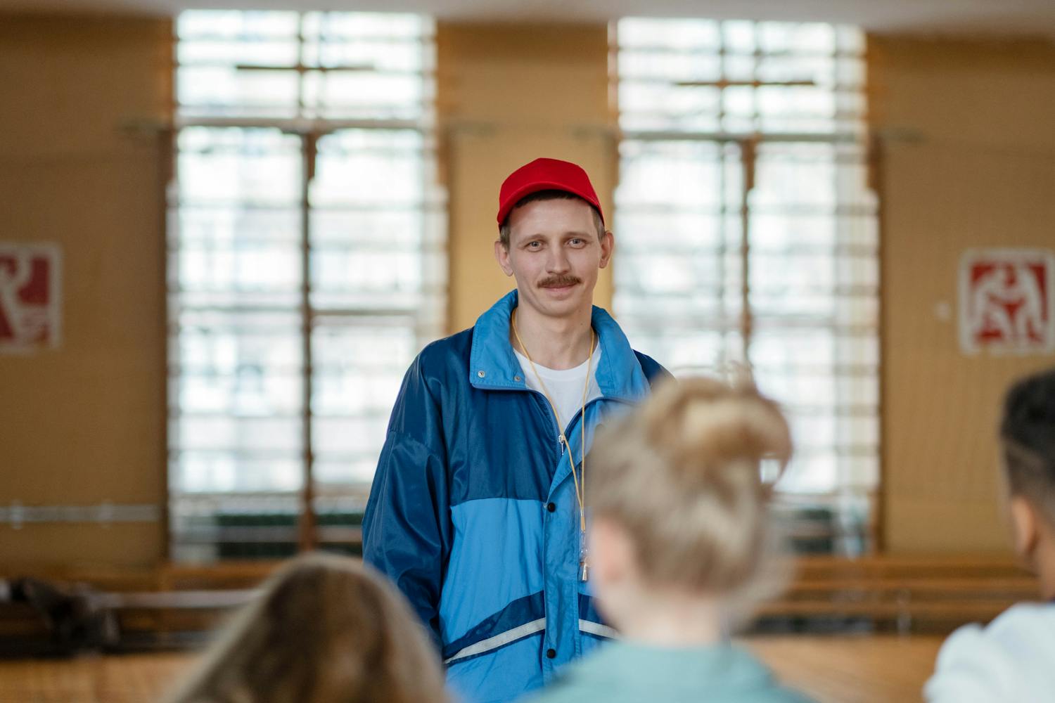 Male coach in a gym engaging with a group of children, wearing casual sportswear.